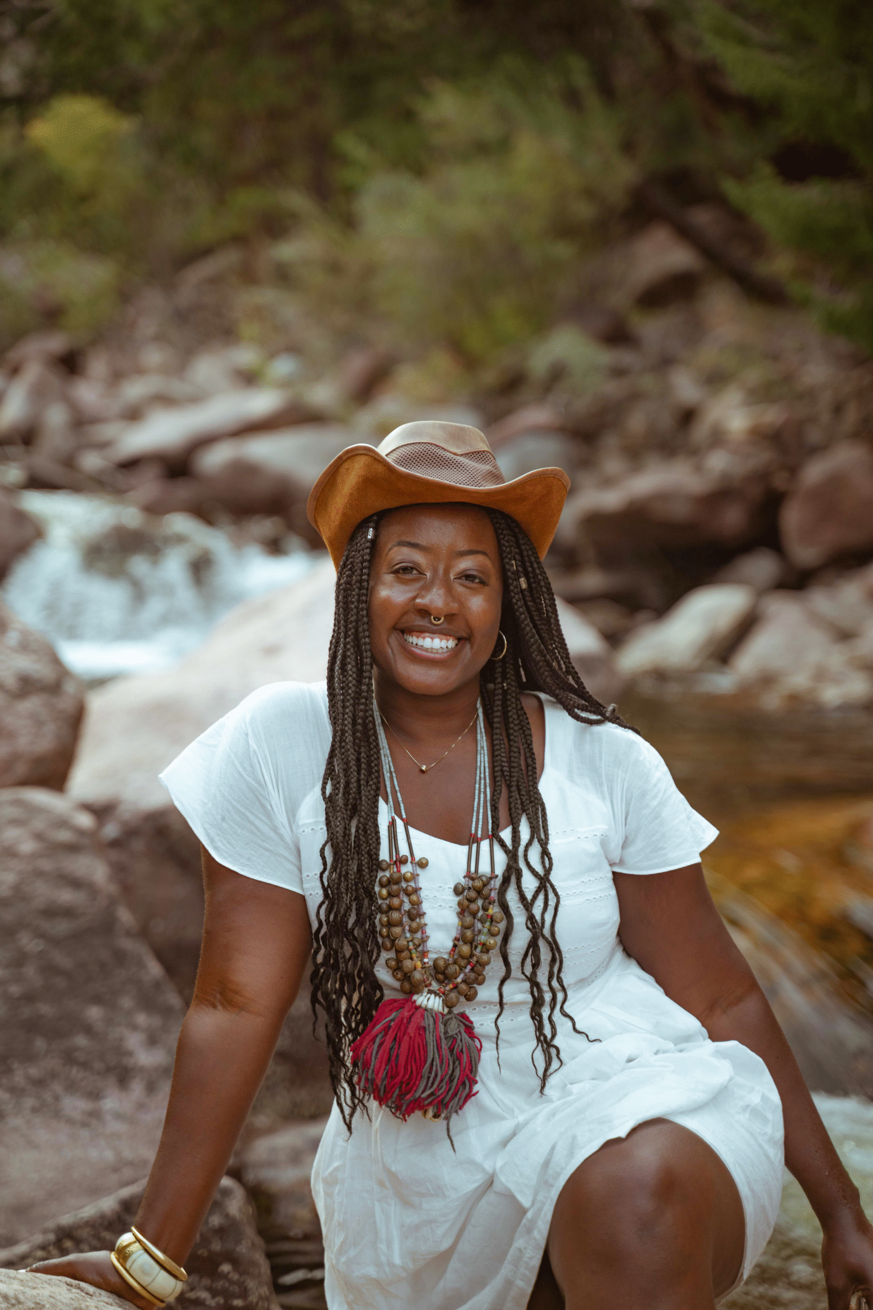 Asia Dorsey smiling while seated on boulders by a river, wearing a white dress and brown hat